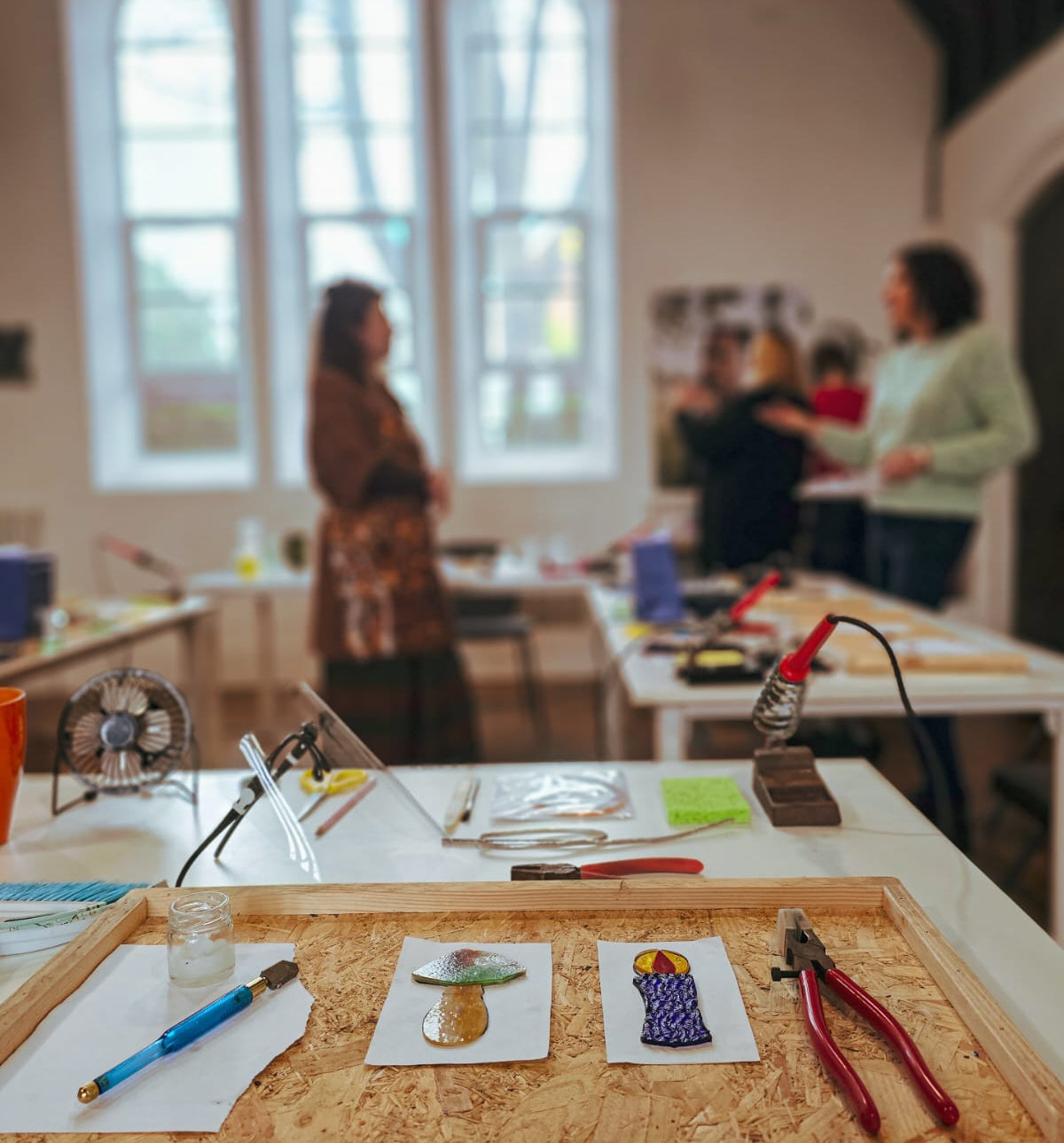 Artistic workspace with tools and half completed stained glass project on a table, blurred background of people and windows.