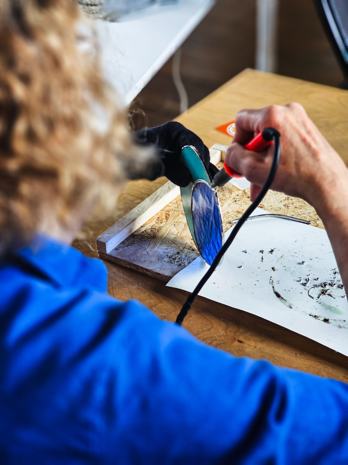 Person using a soldering iron to make a stained glass moon shaped suncatcher on a wooden surface with a blurred background