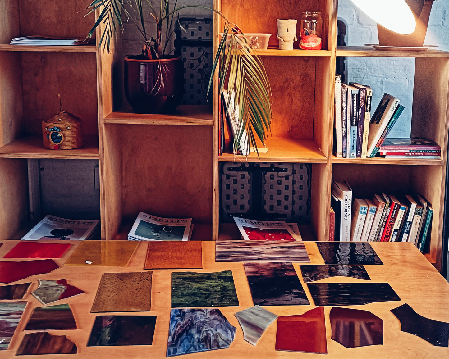 Still life from a stained glass workshop, wooden shelves with books and decorative items, and a table with sheets of coloured glass lay across it.