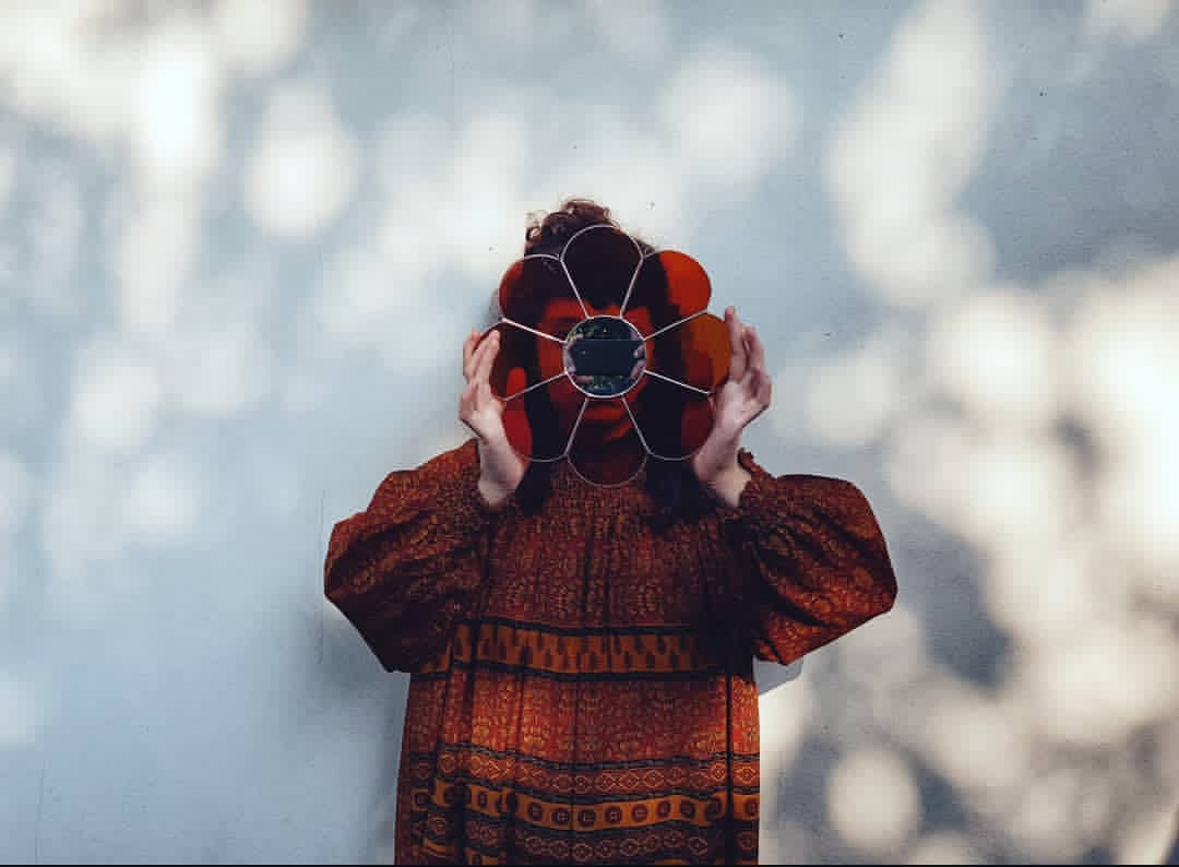 Artist Yiscah Rosemary woman wearing a patterned dress holding a stained glass flower shaped suncatcher in front of their face against a blurred natural background of soft light through leaves