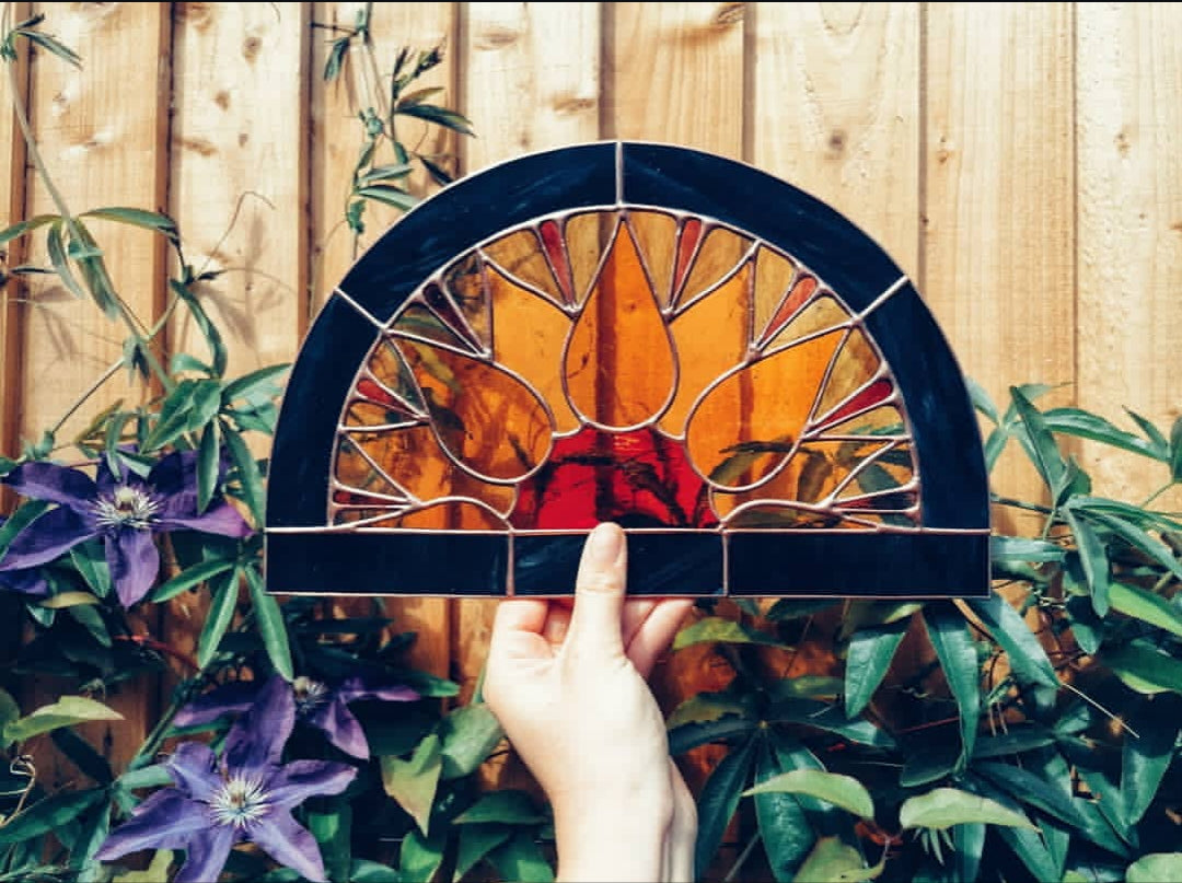 Stained glass panel of a half circle sunflower window design held by a hand with a wooden fence and plants and passionflowers in the background by artist Yiscah Rosemary