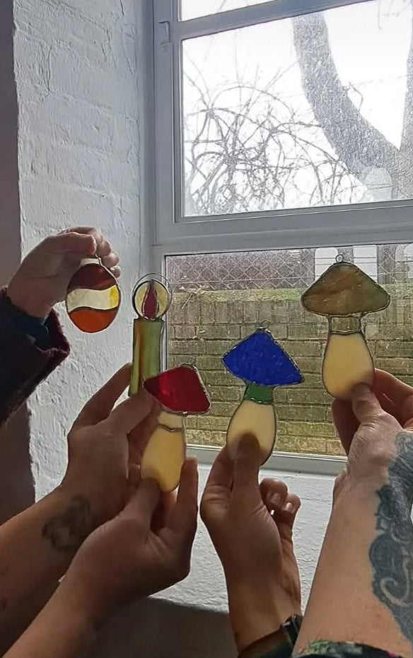 Hands holding colorful stained glass mushroom suncatcher window hangings in front of a window at a workshop.