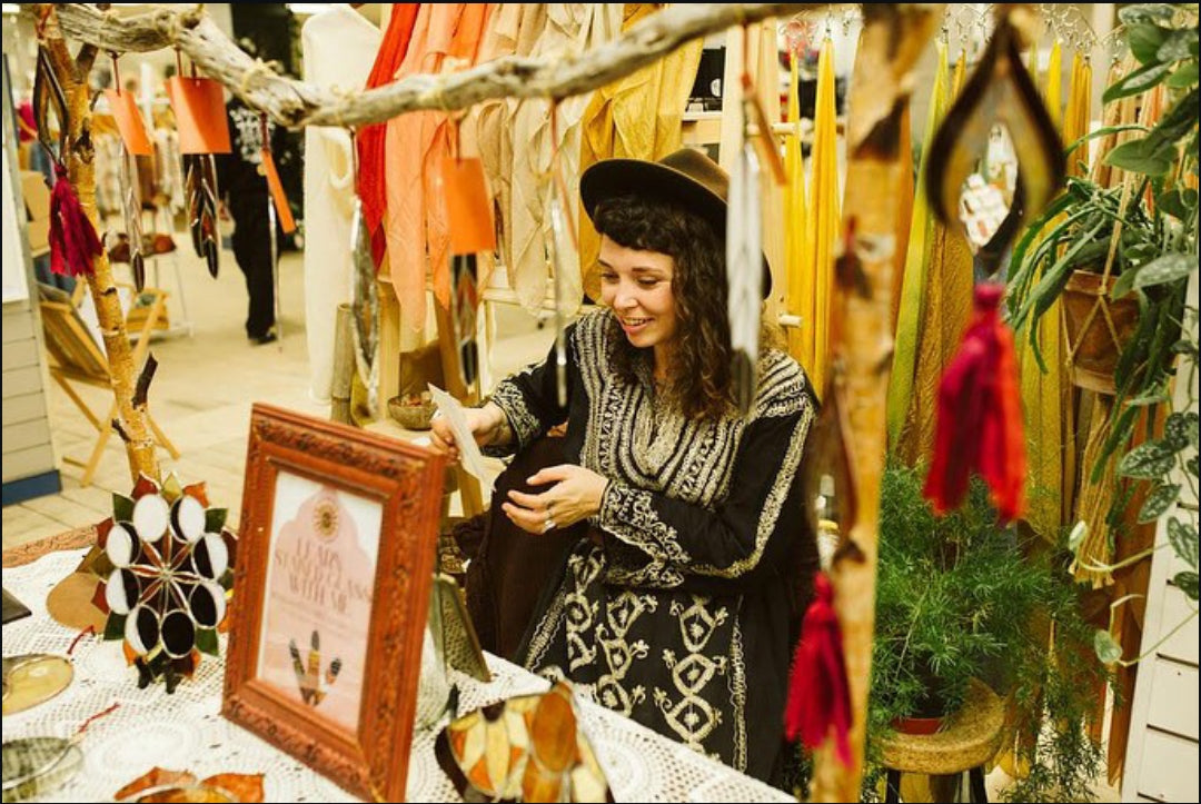 Artist Yiscah Rosemary in an indian embroidered dress and wide brim hat smiling behind her stall of glass art at the Southcoast Makers Market indoor market stall in Bournemouth.