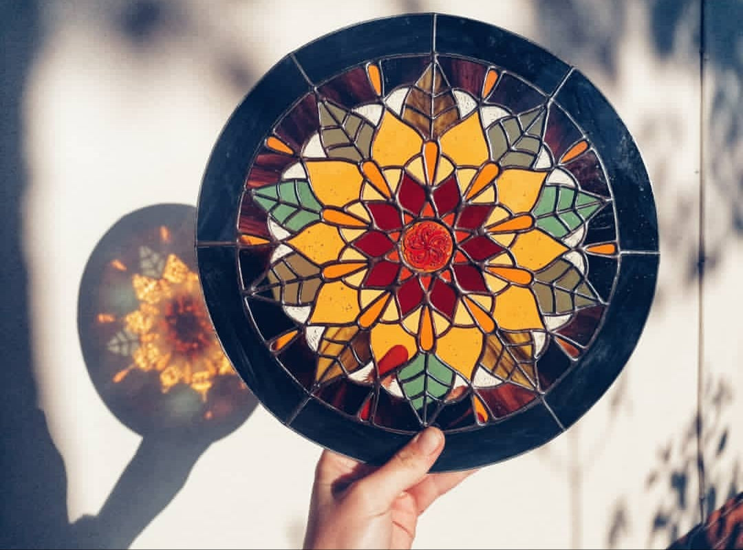Hand holding a round stained glass window with colourful geometric design of a sunflower by artist Yiscah Rosemary.